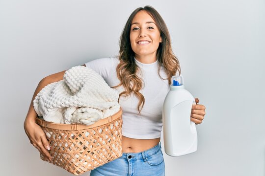Young brunette woman holding laundry basket and detergent bottle smiling with a happy and cool smile on face. showing teeth.