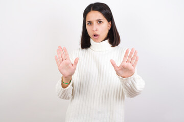 Young brunette woman wearing white knitted sweater against white background Moving away hands palms showing refusal and denial with afraid and disgusting expression. Stop and forbidden.