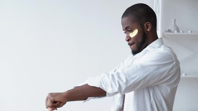 Metrosexual lifestyle. Male grooming. Morning fun. Relaxed confident African man with gold hydrogel eye patches dancing wearing white shirt in light modern room at home.