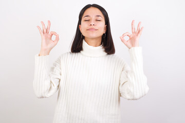 Young brunette woman wearing white knitted sweater against white background relax and smiling with eyes closed doing meditation gesture with fingers. Yoga concept.