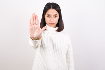 Young brunette woman wearing white knitted sweater against white background doing stop sing with palm of the hand. Warning expression with negative and serious gesture on the face.