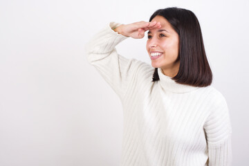 Young brunette woman wearing white knitted sweater against white background very happy and smiling looking far away with hand over head. Searching concept.