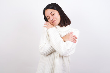 Young brunette woman wearing white knitted sweater against white background Hugging oneself happy...