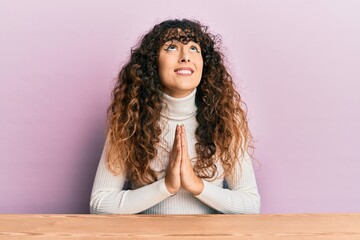Young hispanic girl wearing casual clothes sitting on the table begging and praying with hands...