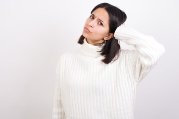 Obraz premium Young brunette woman wearing white knitted sweater against white background confuse and wonder about question. Uncertain with doubt, thinking with hand on head. Pensive concept.