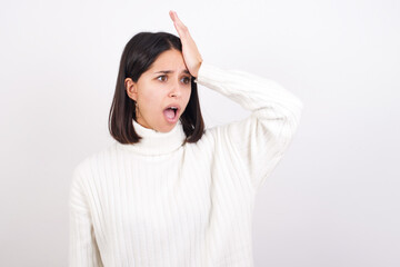 Young brunette woman wearing white knitted sweater against white background surprised with hand on head for mistake, remember error. Forgot, bad memory concept.