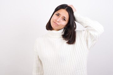 Young brunette woman wearing white knitted sweater against white background being confused and wonders about something. Holding hand on her head, uncertain with doubt. Pensive concept.