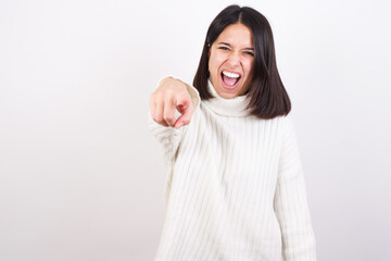Young brunette woman wearing white knitted sweater against white background pointing displeased and frustrated to the camera, angry and furious ready to fight with you.