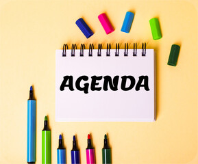 The word AGENDA written in a white notebook on a beige background near multi-colored markers