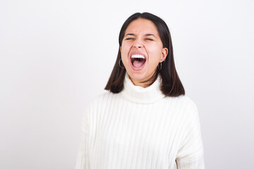 Young brunette woman wearing white knitted sweater against white background angry and mad screaming frustrated and furious, shouting with anger. Rage and aggressive concept.