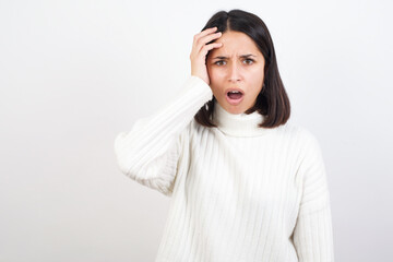 Embarrassed Young brunette woman wearing white knitted sweater against white background with...