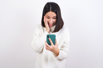 Fototapeta premium Young brunette woman wearing white knitted sweater against white background holding in hands cell reading sms using new app 5g