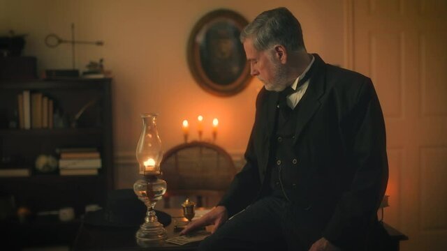 A mature pensive 19th century businessman sitting on the edge of his desk starts counting some money that is lying on the desk then picks up a document and starts reading it.