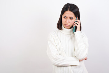 Sad Young brunette woman wearing white knitted sweater against white background  talking on...