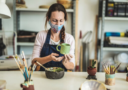 Caucasian woman works with ceramics and paints finished cup while wearing protective face mask for coronavirus