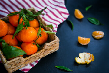 Fresh ripe mandarins, clementine, tangerine with green leaves in the straw bowl on a striped napkin and dark blue stone background. Local citrus fruit harvest. Selective focus.