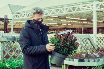 A young man in a protective face mask covering choosing a new green plant in an outdoor garden trading center. Preparing for the new spring planting season. Hobby and leisure time. Selective focus.