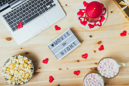 St. Valentine's day Movie night concept. Movie night message on board, laptop, popcorn, red candle, gift, hearts decor, two cups of cocoa with marshmallows for a couple. Cozy holiday plans for lovers.
