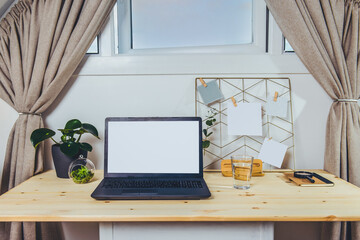 The modern workplace - wooden desk with laptop mockup white empty screen, mood board with pined notes and photo, green plants at work space under a window in home office room interior. Selective focus