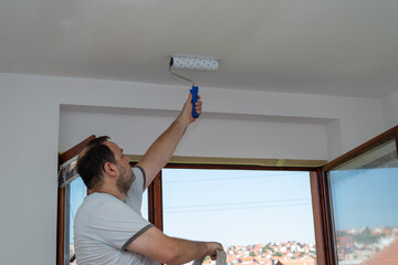 Hard working man painting ceiling with a roller in white. Open window with red roofs is behind him