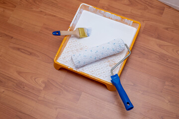 Painting equipment on a floor: a paint tray with white paint, a paint roller and a paintbrush