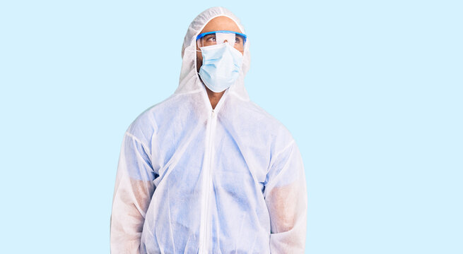 Young hispanic man wearing doctor protection coronavirus uniform and medical mask smiling looking to the side and staring away thinking.