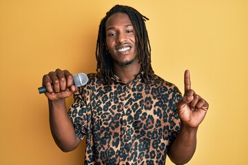 African american man with braids singing song using microphone smiling with an idea or question...