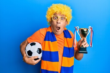 Senior hispanic man football hooligan cheering game holding ball and trophy afraid and shocked with surprise and amazed expression, fear and excited face.