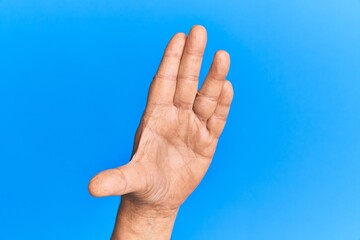 Hand of senior hispanic man over blue isolated background greeting doing vulcan salute, showing hand palm and fingers, freak culture