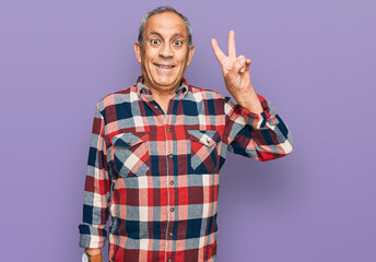 Senior hispanic man wearing casual clothes smiling with happy face winking at the camera doing victory sign. number two.