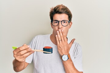 Handsome caucasian man eating sushi using chopsticks covering mouth with hand, shocked and afraid for mistake. surprised expression