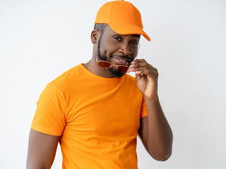 Male fashion. Summer look. Fun lifestyle. Confident happy stylish cool African man model in orange t-shirt matching cap holding sunglasses smiling looking at camera isolated on light background.