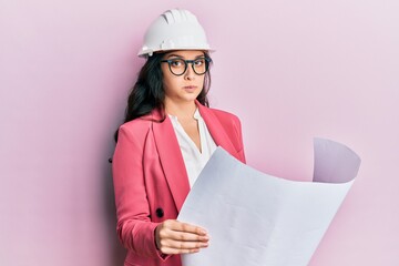 Beautiful middle eastern woman looking at paper blueprints wearing safety helmet puffing cheeks with funny face. mouth inflated with air, catching air.