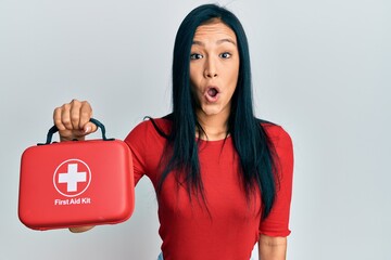 Beautiful hispanic woman holding first aid kit scared and amazed with open mouth for surprise,...