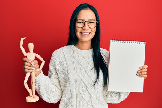 Beautiful hispanic woman holding small wooden manikin and notebook smiling with a happy and cool smile on face. showing teeth.