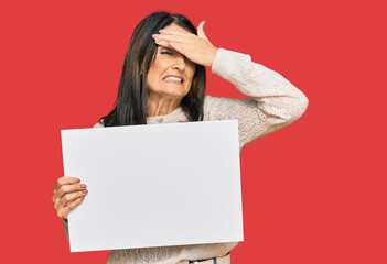 Middle age brunette woman holding blank empty banner stressed and frustrated with hand on head, surprised and angry face