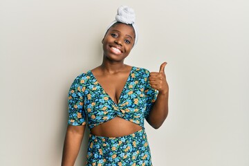 Young african woman wearing hair turban and african style smiling with happy face looking and pointing to the side with thumb up.