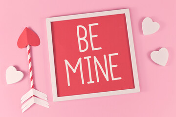 Valentine's Day composition with picture frame with text 'Be Mine', cupid love arrows, and white hearts on pink background