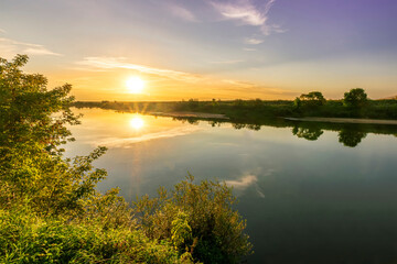 Scenic view at beautiful summer river sunset with reflection on water with green bushes, grass, golden sun rays, calm water ,deep blue cloudy sky and glow on a background, spring evening landscape