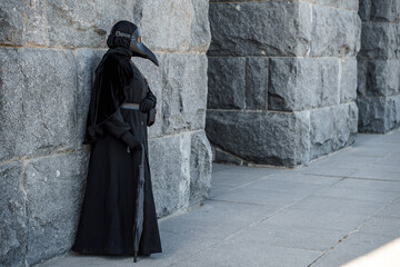 plague doctor stands against a stone wall