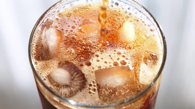 Top view close up footage of a glass with ice being filled with cola, you can see the fresh bubbles of the beverage. Slow motion.