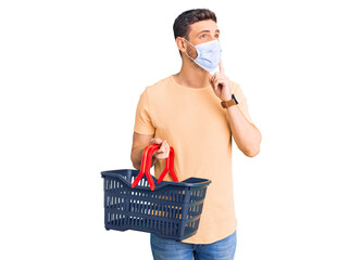 Handsome young man with bear wearing shopping basket and medical mask serious face thinking about...