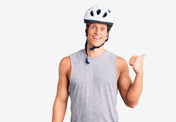 Young handsome man wearing bike helmet smiling with happy face looking and pointing to the side with thumb up.