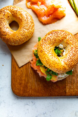 A breakfast composition with a sandwich made of sesame bagel, smoked salmon, white cheese on a cutting board, top view.