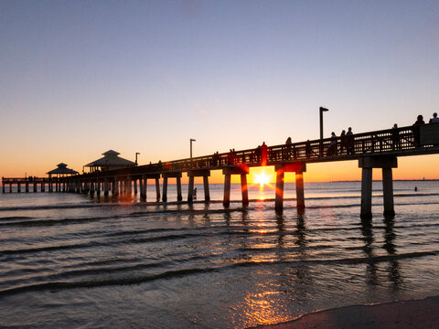 Fototapeta The sun setting below the Ft Myers Beach Pier with the pier and people in silhouette on a blue evening.