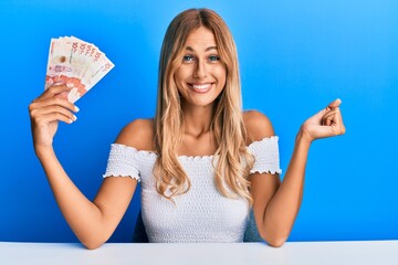 Beautiful blonde young woman holding 10 colombian pesos banknotes screaming proud, celebrating...