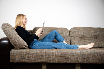 Smiling young caucasian female lying on cozy sofa with tablet watching video movie or shopping online at home