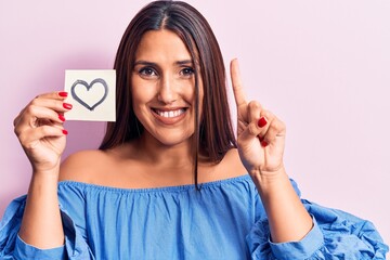 Young beautiful brunette woman holding reminder with heart smiling with an idea or question pointing finger with happy face, number one