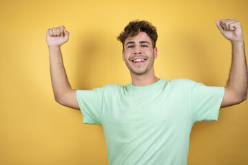 Handsome man wearing a green casual t-shirt over yellow background very happy and excited making winner gesture with raised arms, smiling and screaming for success.