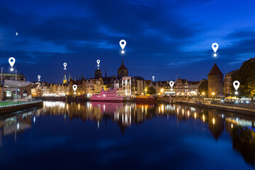 Map pin icons on Gdansk cityscape at dusk. Motlawa River and lit old buildings on the Long Bridge waterfront at the Main Town (Old Town) in Gdansk, Poland, in the evening. Copy space.